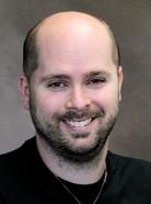 A smiling man with a shaved head, a beard, and wearing a black shirt, is pictured against a neutral background.